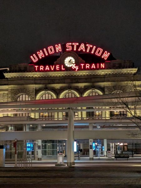 nighttime exterior view of Denver Union Station with a sign reading travel by train