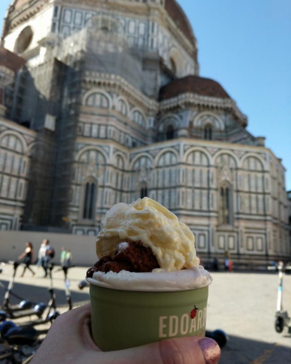 a scoop of gelato in a cup in the foreground of a photo of the exterior of the Duomo from the Piazza del Duomo in Florence, Italy