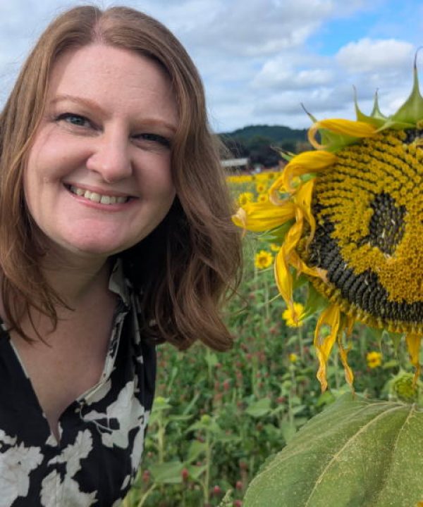 travel agent smiling in a sunflower field next to a sunflower with a smiley face in its center
